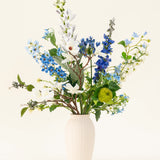 Het Zijden kunstbloemen boeket Cool Breeze, met witte lelies, blauwe ridderspoor en groen gebladerte in een crèmekleurige vaas, steekt af tegen een effen achtergrond voor een verfrissende touch.