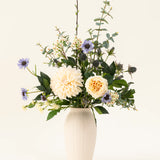 Een witte vaas van keramiek bevat het Zijden kunstbloemen boeket Petal Poetry, met crèmekleurige dahlia's en rozen, lichtpaarse bloemen, groene distels en allerlei groen tegen een effen lichte achtergrond.