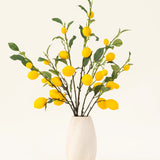 Een vaas met witte structuur toont het Zijden kunstbloemen boeket Lemons, met groene bladeren en levendige gele citroenen tegen een lichte achtergrond.