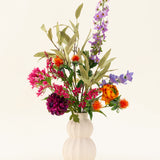Een witte, gestructureerde vaas toont het Zijden kunstbloemen boeket Colourful Svenski, met levendige paarse, oranje, roze en blauwe kunstbloemen met groene bladeren die elke ruimte opfleuren tegen een eenvoudige lichte achtergrond.