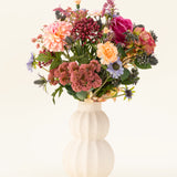 In een witte, gebogen vaas van keramiek staat het Zijden kunstbloemen boeket Pretty Pastel - een kleurrijke mix van duurzame bloemen zoals roze rozen, dahlia's, paarse en oranje bloemen en groen - prachtig gerangschikt tegen een lichte achtergrond.