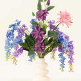 Een witte golvende vaas toont het Zijden kunstbloemen boeket Indigo Dreams - een arrangement van paarse, blauwe en roze kunstbloemen met groene bladeren - tegen een effen lichte achtergrond.