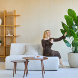 Een vrouw in een bruine jurk zit op een witte bank, glimlacht en houdt een drankje vast terwijl ze de Kunstplant Strelitzia Real Touch Deluxe 180 cm aanraakt in een moderne, minimalistische woonkamer met houten planken en een salontafel.