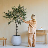 Een vrouw in losse beige kleding staat binnen naast een Kunst Olijfboom van 200 cm en raakt zachtjes de bladeren aan. Vlakbij staan een houten kruk en een consoletafel met boeken en een diffuser tegen de beige muur.