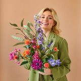 Een lachende blonde persoon in een groen pak houdt het levendige Zijden kunstbloemen boeket Colourful Svenski tegen een effen, lichtbruine achtergrond.