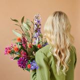 Een persoon met lang, golvend blond haar en een groene jas is van achteren gezien, terwijl hij het Zijden kunstbloemen boeket Colourful Svenski vasthoudt tegen een effen beige achtergrond.