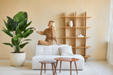 Een vrouw in een bruine jumpsuit zit op de rugleuning van een witte bank en raakt het blad aan van een Kunstplant Strelitzia Real Touch Deluxe 180 cm in een minimalistische woonkamer met houten tafels en planken.