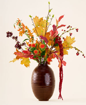 In een bruine vaas staat het boeket ‘Rustic Glow’ van Zijden kunstbloemen, met gele, oranje en rode bladeren, rode bessen, oranje bloemen en lange, hangende rode amaranthus tegen een neutrale achtergrond.