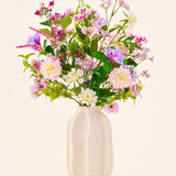In een witte keramische vaas staat een kleurrijk boeket van roze, paarse en witte bloemen uit de collectie ‘Zijden kunstbloemen boeket Royal Bloom’, geaccentueerd met groene bladeren en gepresenteerd tegen een effen, lichte achtergrond.