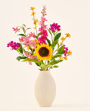 In een beige keramische vaas staat het zijden kunstbloemenboeket ‘Tiny Sunshine’, bestaande uit een kunstboeket van levensechte bloemen, zoals een grote zonnebloem, roze en magenta bloesems en gele bloemen met groen blad, tegen een effen lichte achtergrond. Dit arrangement van Easyplants bestaat uit prachtige zijden bloemen die elke ruimte langdurige schoonheid verlenen.
