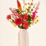 In een witte keramische vaas staat het boeket ‘Summer Blush’ van Zijden kunstbloemen, bestaande uit roze, gele en oranje bloemen met groene bladeren, tegen een effen lichte achtergrond.