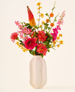 In een witte keramische vaas staat het boeket ‘Summer Blush’ van Zijden kunstbloemen, bestaande uit roze, gele en oranje bloemen met groene bladeren, tegen een effen lichte achtergrond.
