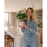 Een glimlachende vrouw in een spijkeroutfit houdt de Kunstplant Gardenia UV 39 cm wit vast in een moderne keuken met houten krukken, witte werkbladen en een neutrale inrichting.