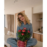 Een vrouw met krullend blond haar, gekleed in een spijkerjumpsuit en roze sneakers, zit in kleermakerszit op een aanrecht en glimlacht terwijl ze de Kunstplant Gardenia UV 39 cm in fuchsia vasthoudt.