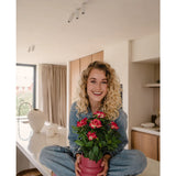 Een glimlachende vrouw met krullend blond haar zit in kleermakerszit op een lichte, moderne aanrecht, met in haar handen de Kunstplant Gardenia UV 39 cm fuchsia – een roze potplant met levendig rode bloemen. Ze draagt een spijkerbroek, een blauw shirt en roze schoenen.
