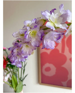 Close-up van de Kunstbloem Delphinium 105 cm paars, geschikt in een vaas, met op de achtergrond een roze en rood bloemenkunstwerk aan de muur.