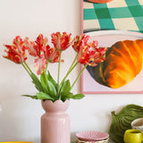 Op een tafel met kleurrijk servies staat een roze vaas met daarin de Kunstbloem Franse Parkiet Tulp 76 cm rood/geel, terwijl op de achtergrond gedeeltelijk een ingelijst schilderij van een croissant en een kopje te zien is.