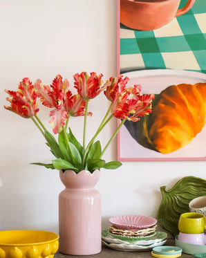 Op een tafel met kleurrijk servies staat een roze vaas met daarin de Kunstbloem Franse Parkiet Tulp 76 cm rood/geel, terwijl op de achtergrond gedeeltelijk een ingelijst schilderij van een croissant en een kopje te zien is.