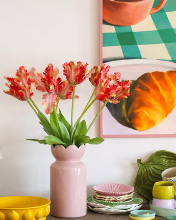 Op een tafel met kleurrijk servies staat een roze vaas met daarin de Kunstbloem Franse Parkiet Tulp 76 cm rood/geel, terwijl op de achtergrond gedeeltelijk een ingelijst schilderij van een croissant en een kopje te zien is.