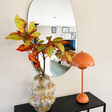 In een golvende spiegel boven een zwarte consoletafel staan een glazen vaas met Kunsttak Kastanje 91 cm rood/groen en een oranje paddestoellamp. De spiegel weerspiegelt een woonkamer met ingelijste kunst aan de muur.