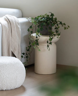 Een Kunst Klimop Deluxe Groen 90 cm staat in een beige vaas op een houten vloer naast een lichtgrijze bank waarop een crèmekleurige plaid ligt en een witte poef met structuur.