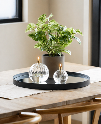 Een Kunst Goudrank groen-wit in pot UV 35 cm staat samen met twee glazen olielampen op een rond spiegelplateau op een houten tafel, verlicht door natuurlijk licht dat door een raam in de buurt naar binnen valt in deze moderne, minimalistische omgeving.