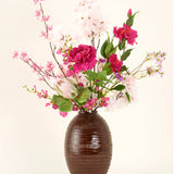 In een vaas met bruine structuur staat het Zijden kunstbloemen boeket Pink Reverie, met roze, magenta en witte kunstbloemen met groene bladeren tegen een effen lichte achtergrond.