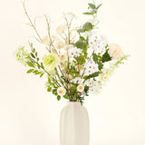 Het Zijden kunstbloemen boeket Claire de Lune heeft delicate witte, lichtgele en groene bloemen in een witte vaas van keramiek, geaccentueerd door weelderig groen tegen een lichte achtergrond.