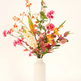 In een vaas met witte structuur staat het Zijden kunstbloemen boeket Wild Peach met kleurrijke kunstbloemen - roze, oranje, geel en wit - en groene en roodachtige bladeren tegen een effen lichte achtergrond.