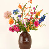 In een vaas met bruine structuur staat het Zijden kunstbloemen boeket Tropical Firework - een arrangement van oranje, roze, paarse, gele en blauwe zijden bloemen met groene bladeren tegen een effen lichte achtergrond.