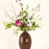 Een Zijden kunstbloemen boeket Serene Bloomfield, met een grote roze protea, witte rozen, paarse en witte bloesems en groen gebladerte, staat in een bruine geribbelde vaas tegen een effen lichte achtergrond.