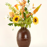 Een vaas met bruine structuur toont het Zijden kunstbloemen boeket Amber Light, met zonnebloemen, witte en oranje bloemen en groen gebladerte tegen een effen achtergrond.