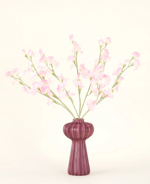 Het Zijden kunstbloemen boeket Erwt lichtroze, met meerdere lichtroze erwt stelen in een geribbelde bordeauxrode vaas, staat tegen een bleke achtergrond - ideaal voor elk kunstbloemen interieur.