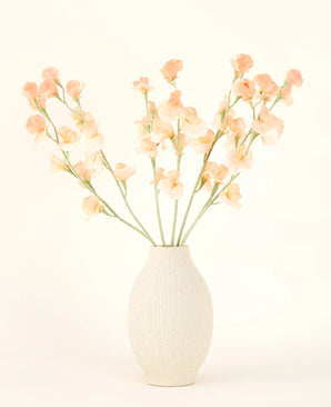 De crèmekleurige vaas van keramiek bevat het Zijden kunstbloemen boeket Erwt perzik, met delicate perzik- en lichtroze bloemen met lange groene stelen tegen een lichte, neutrale achtergrond.