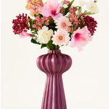 Een boeket kunstbloemen van Zijden, ‘Bloom & Berry’, met roze, witte en rode kunstbloemen, is prachtig geschikt in een hoge, geribbelde, paarse vaas tegen een effen witte achtergrond.