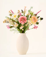 In een witte keramische vaas staat het boeket ‘Backyard Blossom’ van Zijden kunstbloemen, bestaande uit een prachtig arrangement van roze rozen, witte madeliefjes en kleine gele bloemetjes, vervaardigd uit zijden bloemen, aangevuld met weelderig groen blad tegen een effen lichte achtergrond.
