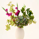 In een witte vaas met structuur staat een boeket van groene hortensia’s, bladrijke takken en felmagenta zijden bloemen tegen een effen lichte achtergrond, waardoor een opvallend ‘Cottage Garden’-boeket van zijden kunstbloemen ontstaat dat elke ruimte een blijvende charme geeft.