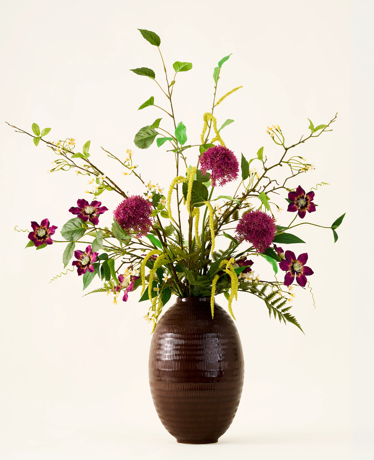 In een bruine, geribbelde vaas staat het boeket „Woodland Charm“ van Zijden kunstbloemen, met paarse alliums, donkerroze bloemen, gele takjes en groen blad tegen een effen, lichte achtergrond.