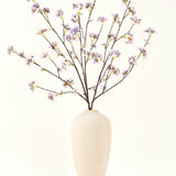 Een hoge, crèmekleurige vaas van keramiek bevat een aantal dunne takken met de tekst Zijden kunstbloemen boeket Kunstbloementak paars, tegen een effen, lichte achtergrond.
