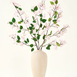 Een beige vaas van keramiek bevat verschillende lange takken met het Zijden kunstbloemen boeket Perzikbloesem roze en groene bladeren, tegen een effen witte achtergrond - perfect voor een kunstbloemen interieur.