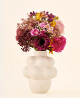 Een boeket van Zijden kunstbloemen, A Touch of Ashley, in roze, paarse en gele tinten, gepresenteerd in een off-white vaas met bubbelvorm en een gestructureerd oppervlak.