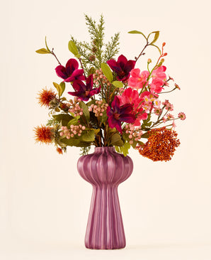 Zijden kunstbloemenboeket Cinnamon Ruby in tinten rood, roze en groen, gepresenteerd in een geribbelde, paarse vaas.