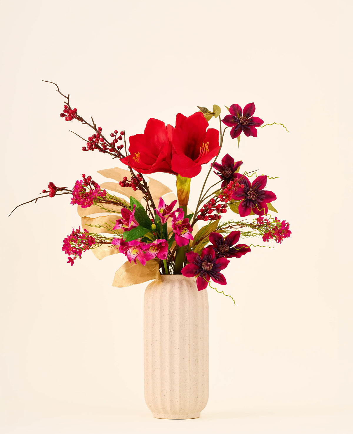 Zijden kunstbloemenboeket Rosy Noël met grote rode amaryllissen, donkerpaarse bloemen en gouden bladeren in een geribbelde, crèmekleurige vaas.