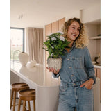 Een glimlachende vrouw in een spijkerjumpsuit staat in een moderne keuken, met de Kunstplant Gardenia UV 39 cm wit in haar handen. Langs een wit aanrecht staan houten krukken opgesteld, en door een raam op de achtergrond stroomt zonlicht naar binnen.
