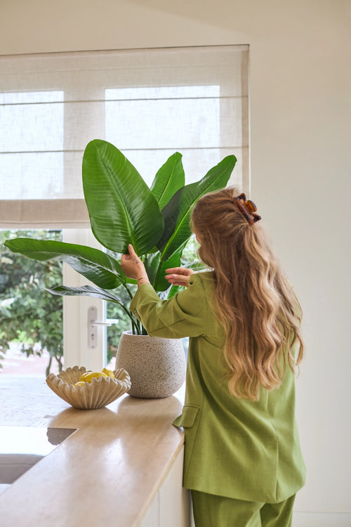 Een vrouw met lang golvend haar, gekleed in een groen pak, verzorgt een Kunstplant Strelitzia Real Touch Deluxe van 90 cm op een toonbank naast een schaal met citroenen in een lichte kamer met een raam.