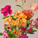 Een levendig boeket kunstbloemen van Zijden, ‘Magnus di Lambo’, bestaande uit een mix van oranje, roze en magenta kunstbloemen, is in een witte vaas geschikt tegen een neutrale achtergrond.