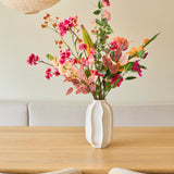 Op een lichte houten tafel staat een witte vaas met een boeket „Zijden kunstbloemen Wild Peach”, samengesteld uit zomerse bloemen. Twee lichte houten stoelen en grote papieren hanglampen maken het minimalistische, gezellige interieur compleet.
