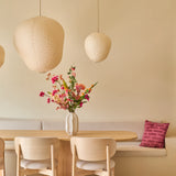 Een minimalistische eethoek met een tafel en stoelen van licht hout, beige muren, drie hanglampen in de vorm van papieren lantaarns, een vaas met het boeket ‘Wild Peach’ van Zijden kunstbloemen dat voor een levendig kleuraccent zorgt, en een ingebouwde bank met daarop een decoratief roze kussen.