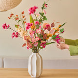 Iemand met een groene mouw schikt het boeket ‘Zijden kunstbloemen Wild Peach’, met kleurrijke zomerse bloemen, in een witte vaas op een lichte houten tafel, met papieren lantaarns en een beige bank op de achtergrond.