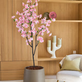 Een Kunst Kersenbloesem 180 cm roze, een potboom met roze bloesems, staat naast een witte stoel met kussens in een moderne kamer met houten planken en decoratieve voorwerpen.